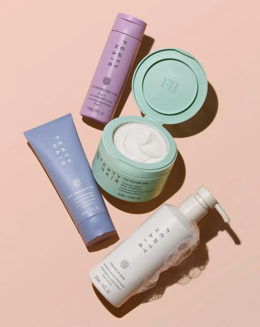 Fenty Hair products flat lay. Includes shampoo, conditioner, hair mask, and heat protectant spray. Pale pink backdrop. Beauty product photography.