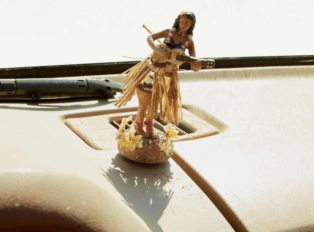 Hawaii hula doll playing a ukulele, wearing a grass skirt and flowers on a car dashboard, sunny lighting, vacation vibes.