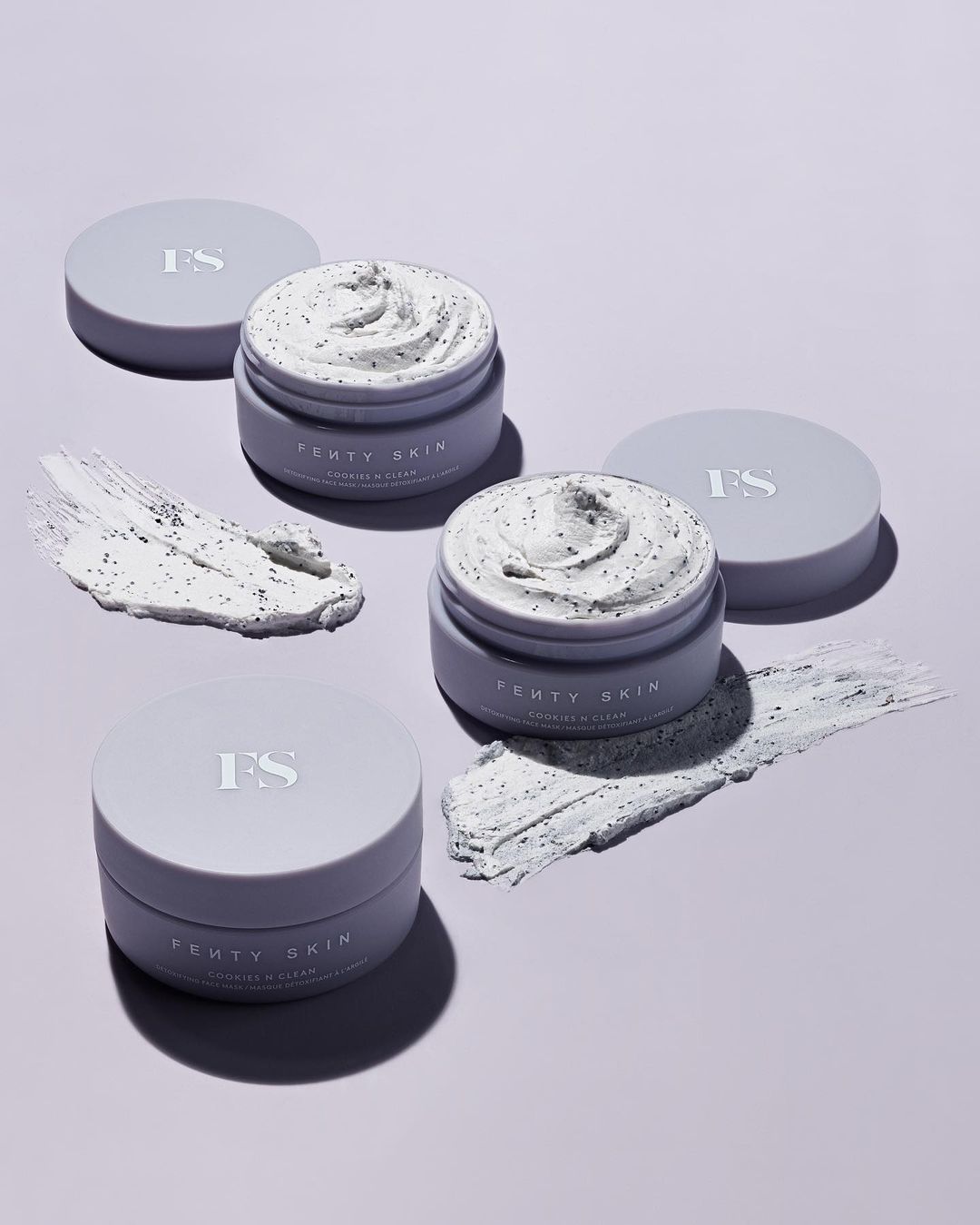 Three open jars of Fenty Skin Coconut & Clean cleansing balm with lids, along with two smears of the balm, on a light purple background.
