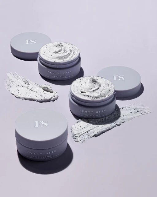 Three open jars of Fenty Skin Coconut & Clean cleansing balm with lids, along with two smears of the balm, on a light purple background.