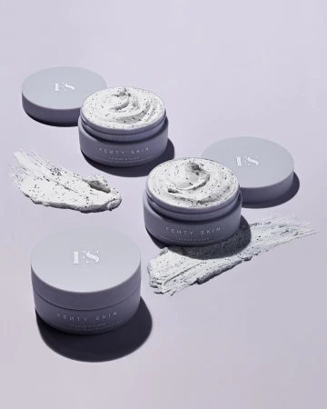 Three open jars of Fenty Skin Coconut & Clean cleansing balm with lids, along with two smears of the balm, on a light purple background.