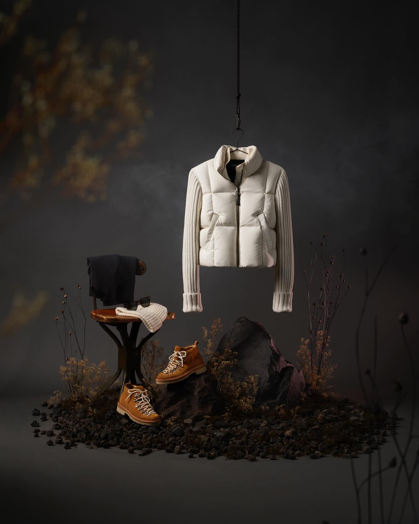 Studio shot of an off-white puffer jacket hanging above brown hiking boots, placed on rocky terrain against a dark backdrop with dried floral elements.