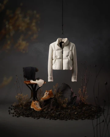 Studio shot of an off-white puffer jacket hanging above brown hiking boots, placed on rocky terrain against a dark backdrop with dried floral elements.