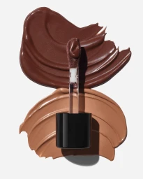 Lip gloss applicator wand resting vertically over two distinct brown and tan cosmetic swatches against a white background.