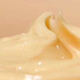 Macro view of pale yellow cosmetic cream swirled into a glossy peak against a solid beige background