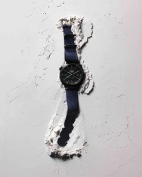 Black wristwatch with a blue fabric strap embedded vertically in a cracked concrete wall.