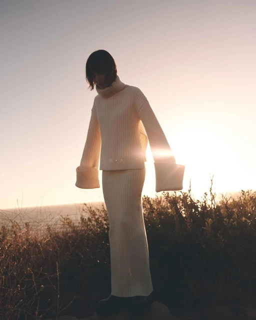 Woman in a cream ribbed turtleneck and skirt poses outdoors at sunset, overlooking the ocean with vegetation in the foreground.