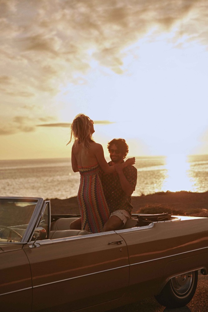 Woman stands in a convertible car with a man, ocean and golden sky at sunset, evoking carefree travel and a retro automotive style.