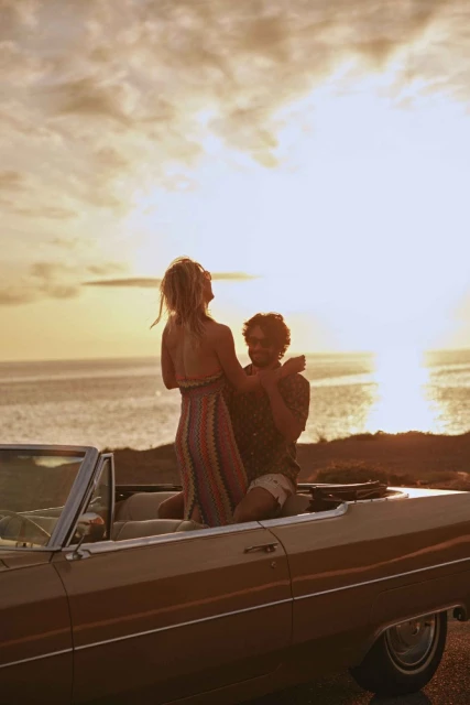 Woman stands in a convertible car with a man, ocean and golden sky at sunset, evoking carefree travel and a retro automotive style.