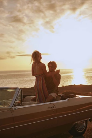 Woman stands in a convertible car with a man, ocean and golden sky at sunset, evoking carefree travel and a retro automotive style.