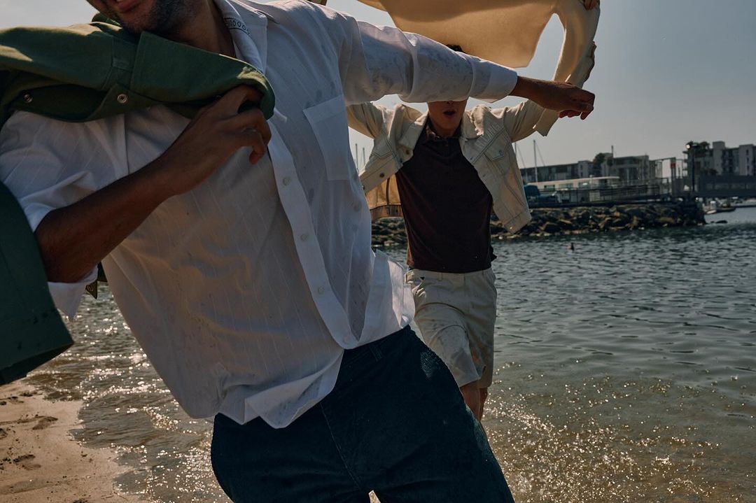 Two men in white shirts walking in shallow water along a beach with distant buildings under a blue sky on a bright sunny day.