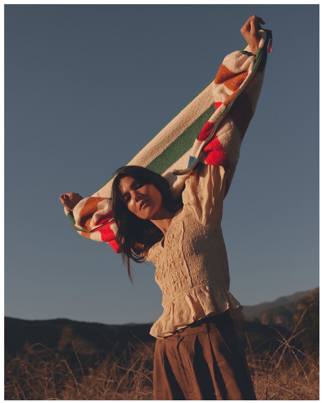 A woman stands in a field, holding a striped blanket overhead against a clear blue sky. She wears a cream-colored blouse and brown pants.