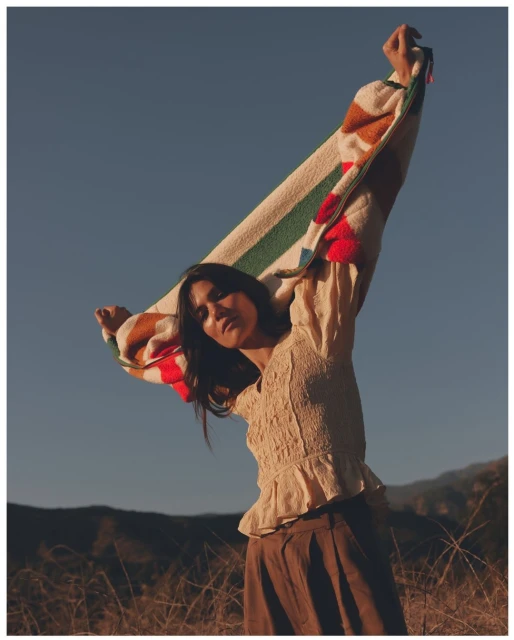 A woman stands in a field, holding a striped blanket overhead against a clear blue sky. She wears a cream-colored blouse and brown pants.