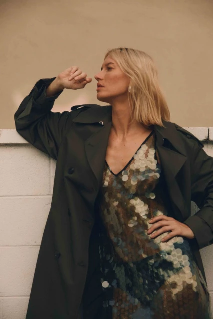 A blonde woman in a trench coat and sequin dress poses against a wall, looking away from the camera in a fashion shot.
