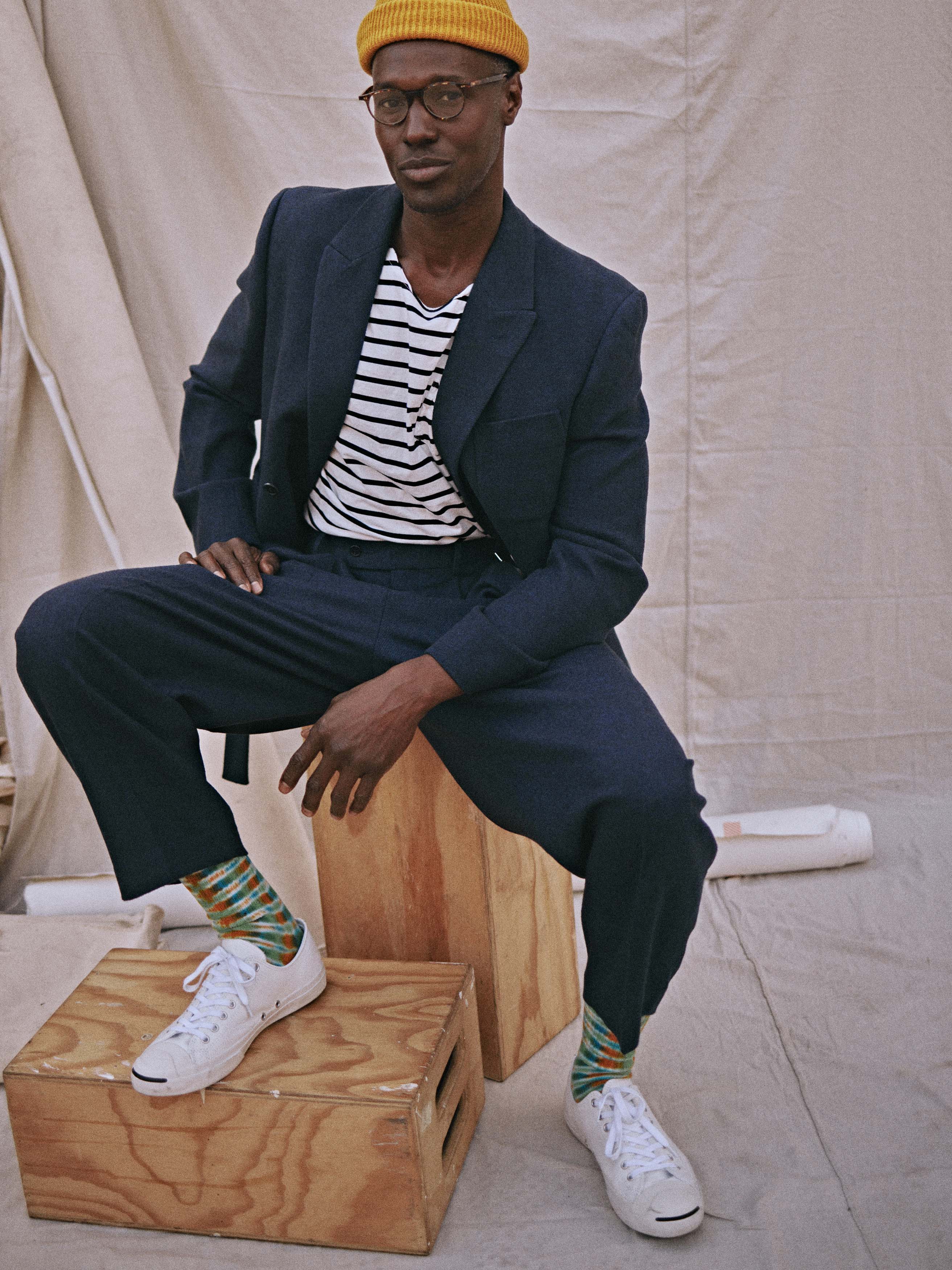 Stylish adult male sits on a wooden crate wearing a navy suit, striped shirt, yellow beanie, glasses, and white sneakers in a neutral indoor setting.