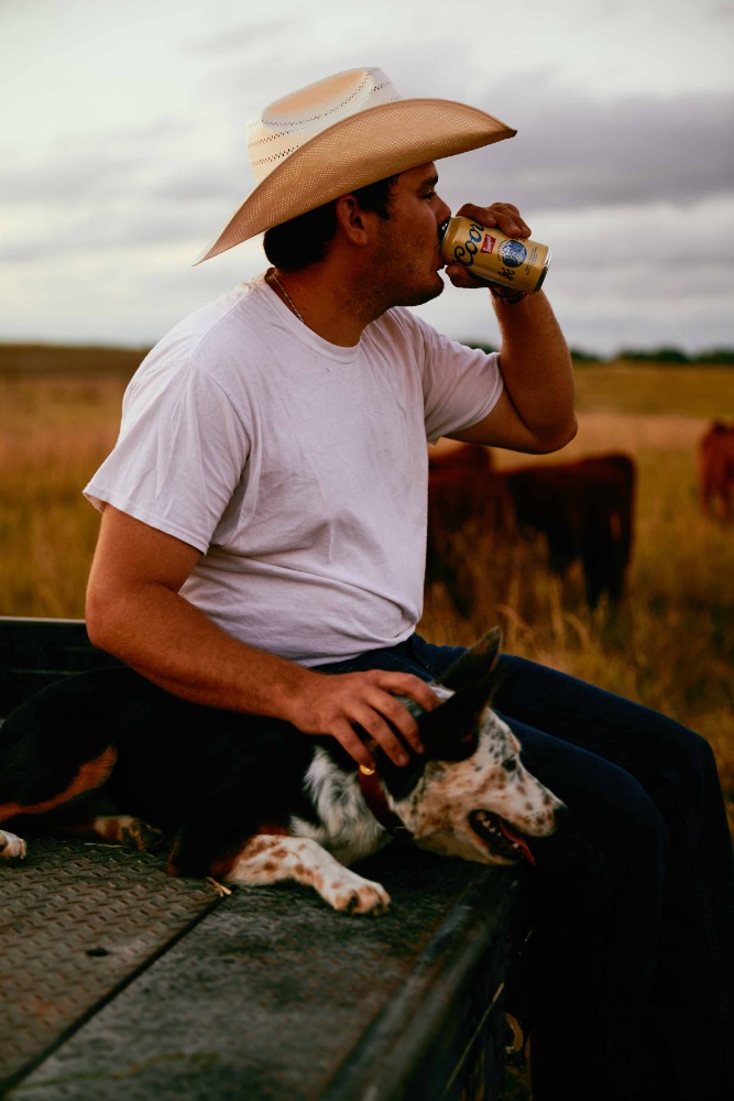 Man wearing a cowboy hat and white t-shirt sitting on a truck tail bed drinking from a can with a dog resting on his lap.