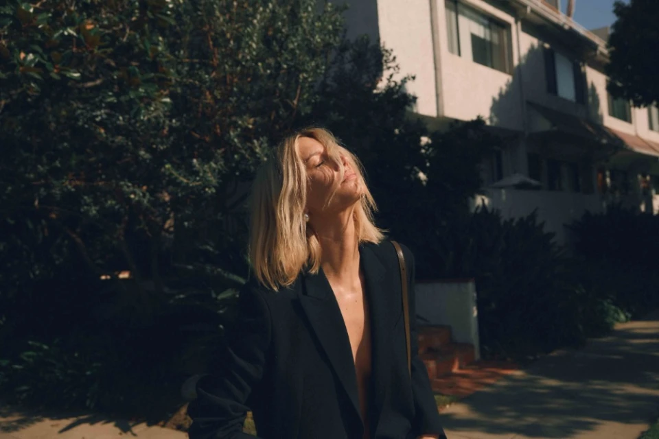 A blonde woman in a black blazer standing outside on a sidewalk with trees and buildings in the background, bathed in sunlight.