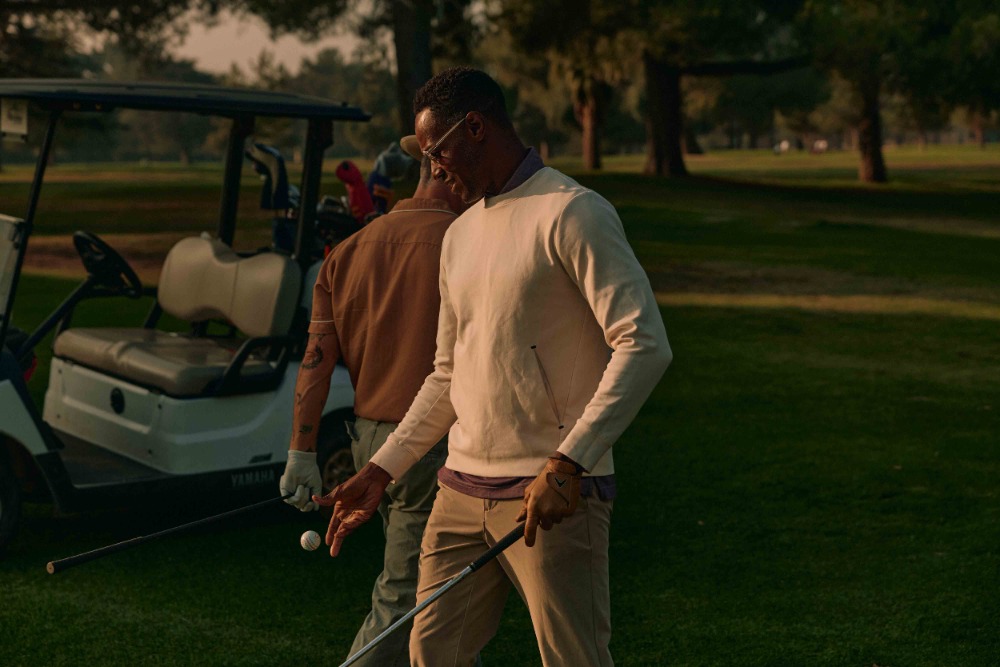 Two adult men play golf on a golf course. One man holds a golf club and golf ball. A golf cart is parked nearby on the green grass.