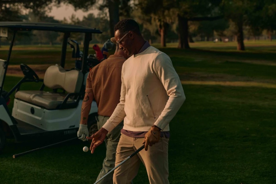 Two adult men play golf on a golf course. One man holds a golf club and golf ball. A golf cart is parked nearby on the green grass.