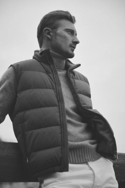 Monochrome portrait of an adult male wearing a puffer vest and turtleneck sweater, leaning on a wooden fence outdoors on an overcast day.