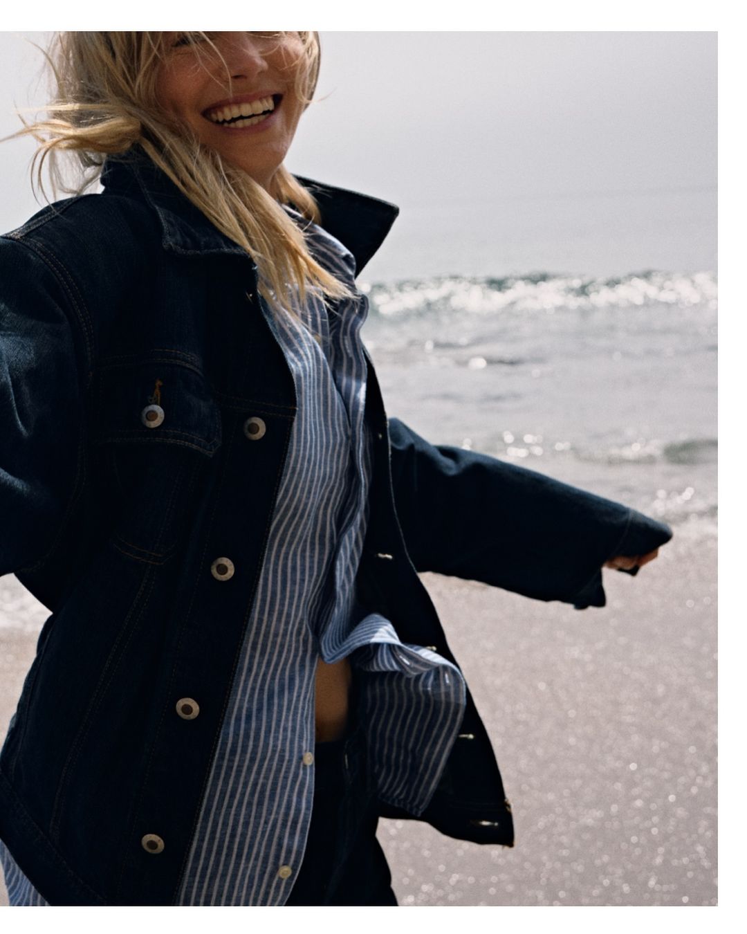 Smiling blonde woman wearing a dark denim jacket and a blue striped shirt walking near the ocean by John Schell