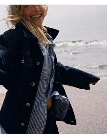 Smiling blonde woman wearing a dark denim jacket and a blue striped shirt walking near the ocean by John Schell