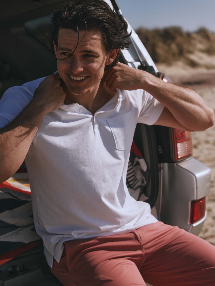 Smiling man in a white shirt leans against a silver SUV on a sandy beach with a dune backdrop on a sunny day.