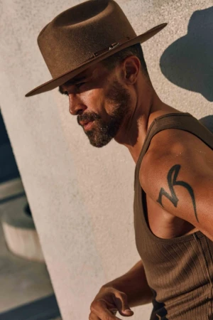 Man wearing a brown fedora hat and olive green tank top leaning against a textured concrete wall in Los Angeles.