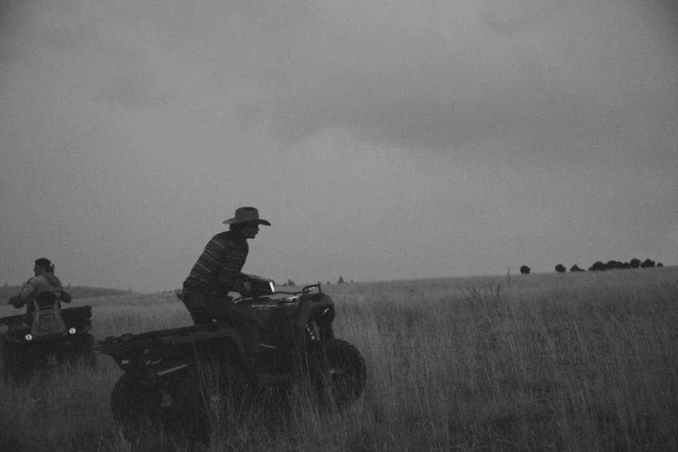 Monochrome shot of two adults riding quad bikes across a grassy field under a cloudy sky, one wearing a cowboy hat.