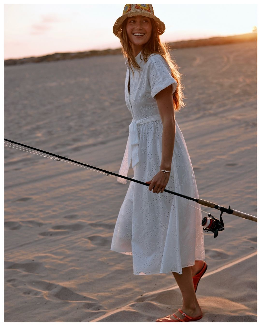 Woman in white dress and hat on beach holding fishing rod, looking happy and smiling against backdrop of sand and sunset.