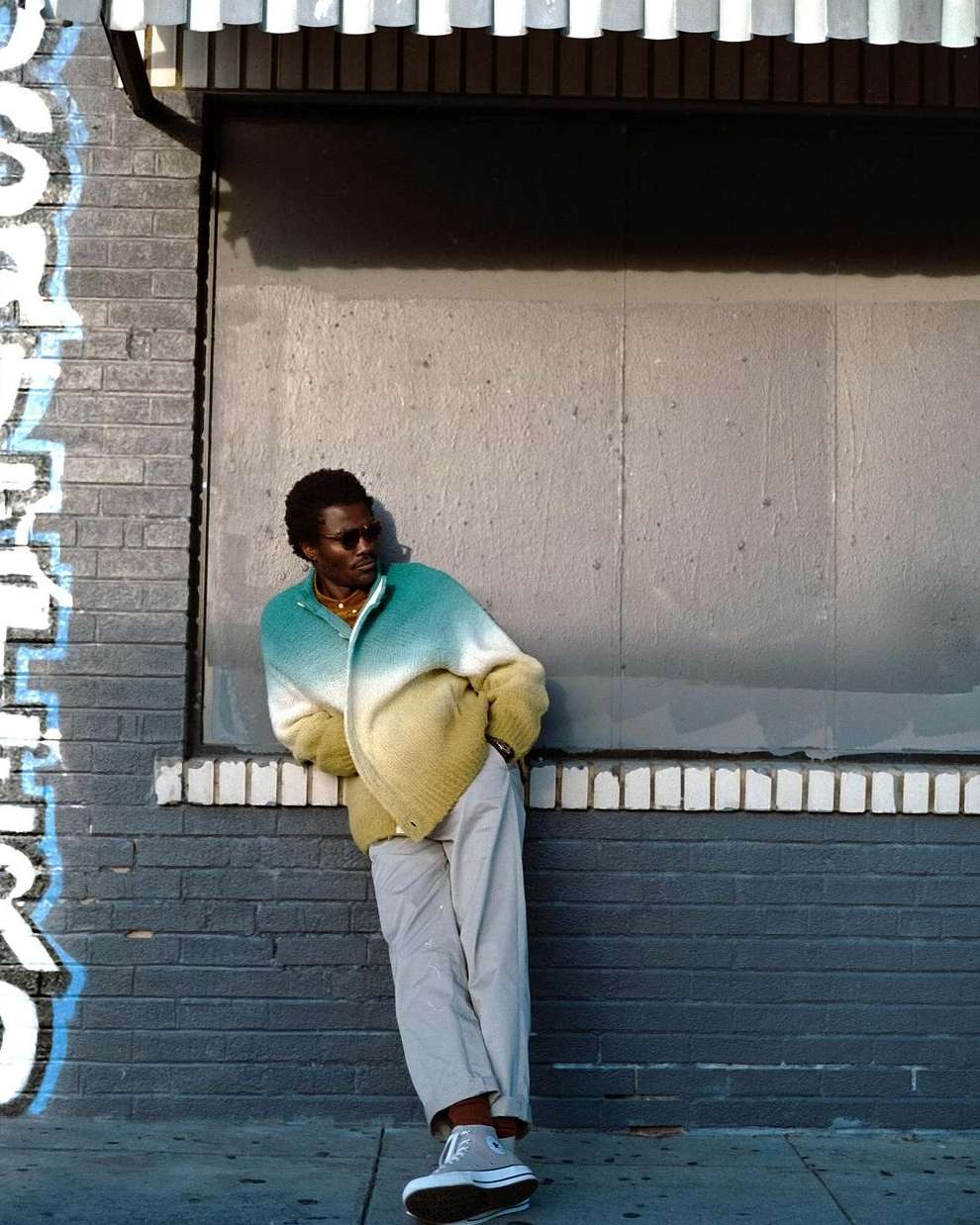 Black man wearing sunglasses and an ombre green and yellow cardigan leaning against a grey brick wall outdoors.