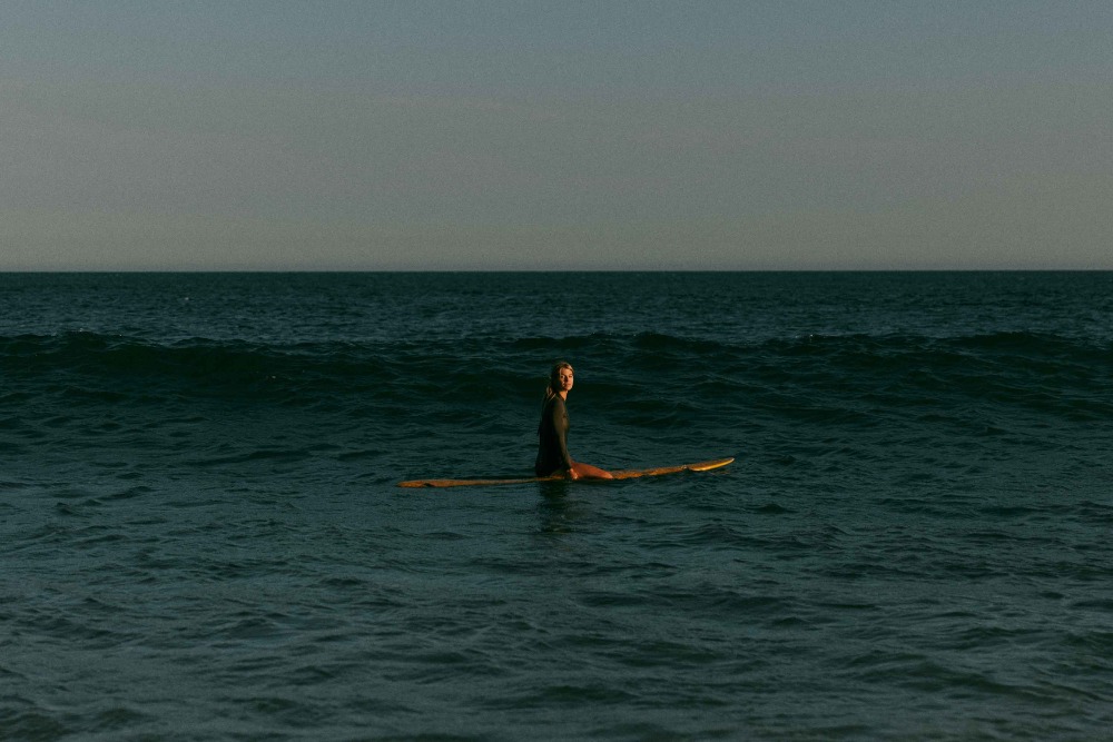 Female surfer sitting on a longboard in the ocean, looking at the camera, with dark teal water and a pale sky in the background.