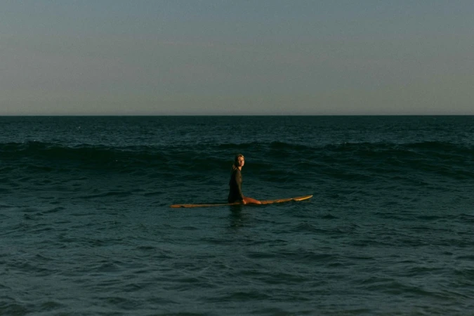 Female surfer sitting on a longboard in the ocean, looking at the camera, with dark teal water and a pale sky in the background.