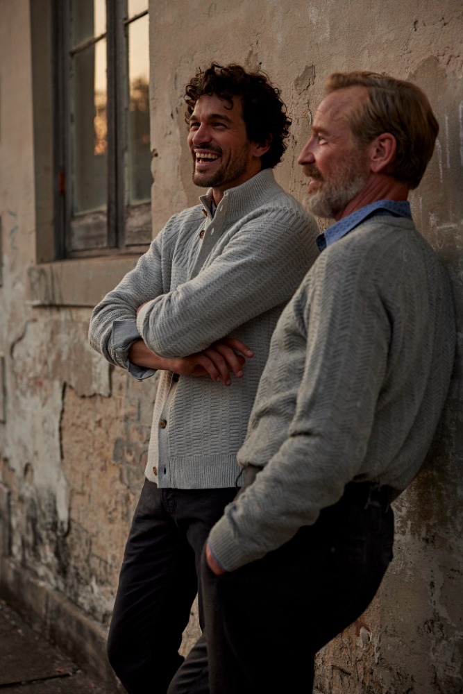 Two men in grey knit cardigans leaning against a weathered stone wall outdoors, laughing together.