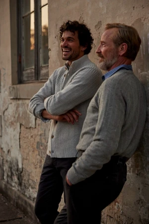 Two men in grey knit cardigans leaning against a weathered stone wall outdoors, laughing together.