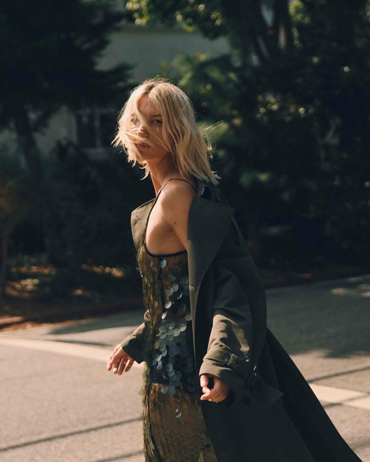 Woman in an olive green dress and trench coat walking outdoors on a street with trees in the background, mid-length shot.