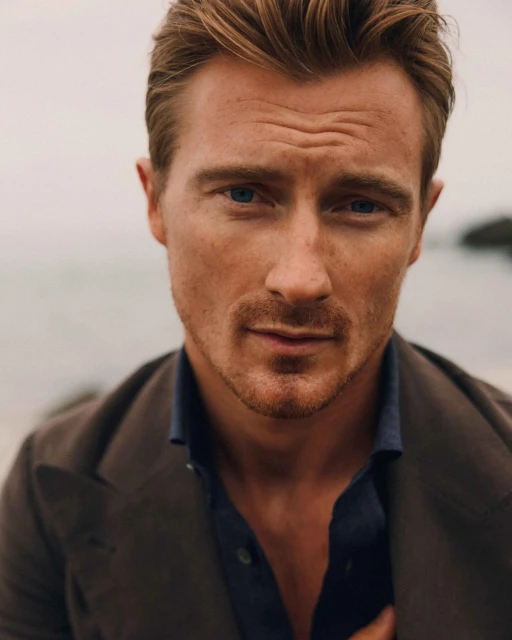 Close-up of a fair-skinned adult man with blue eyes and light brown hair in a brown jacket against a muted outdoor backdrop.