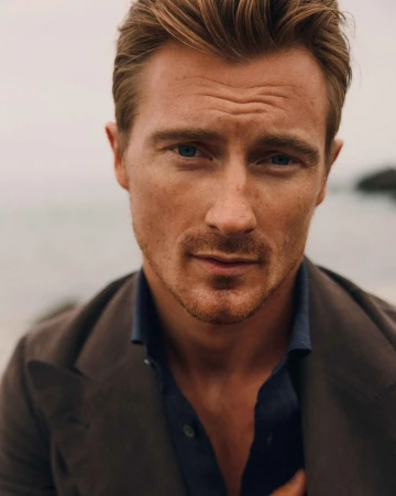 Close-up of a fair-skinned adult man with blue eyes and light brown hair in a brown jacket against a muted outdoor backdrop.