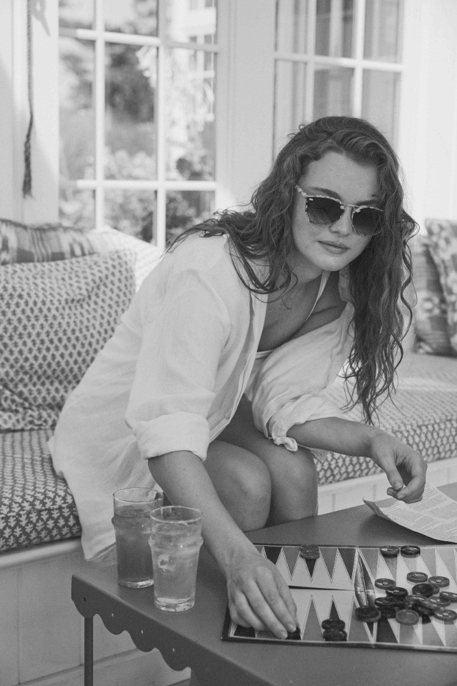 Woman playing backgammon indoors, wearing a white shirt and sunglasses, with drinks on the table; neutral color palette, lifestyle.