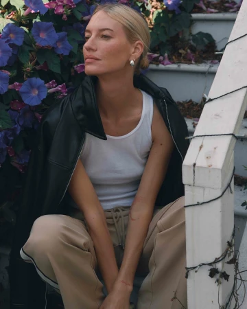 Blonde woman wearing white tank top and black leather jacket sitting on outdoor stairs beside purple flowers, photographed by John Schell.