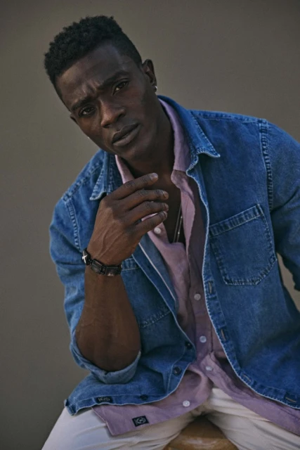 Portrait of a Black man in a denim shirt and white pants sitting and posing against a plain background in a medium shot.