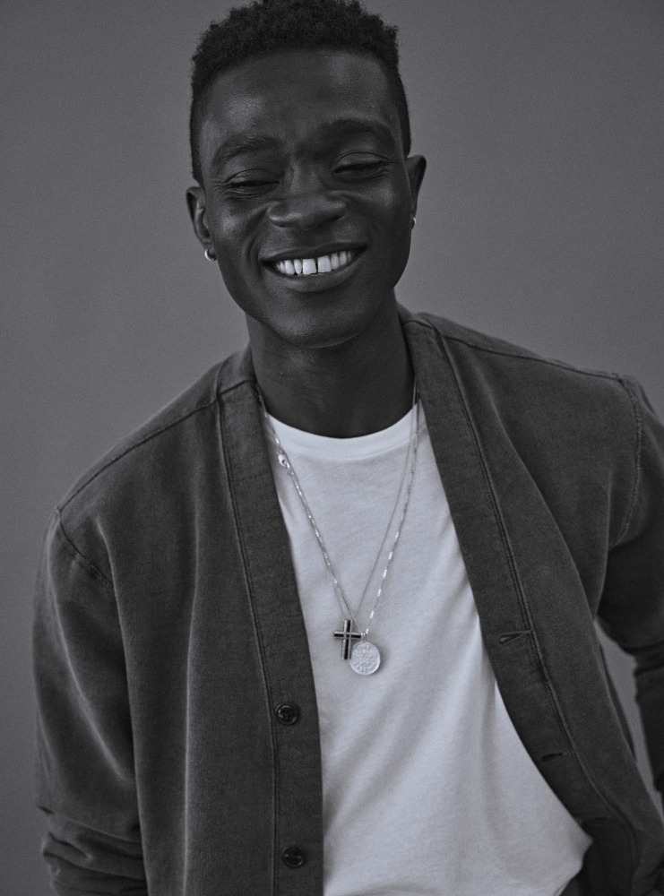 Smiling African American man in casual cardigan and t-shirt, accessorized with silver necklaces, against a solid grey background.
