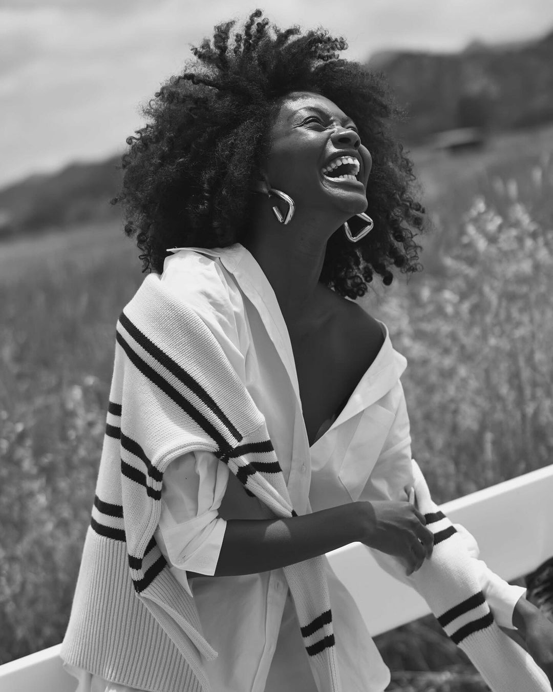 Smiling Black woman in off-shoulder white sweater in a field, silver earrings, monochrome. Outdoor fashion shot of a woman in nature.