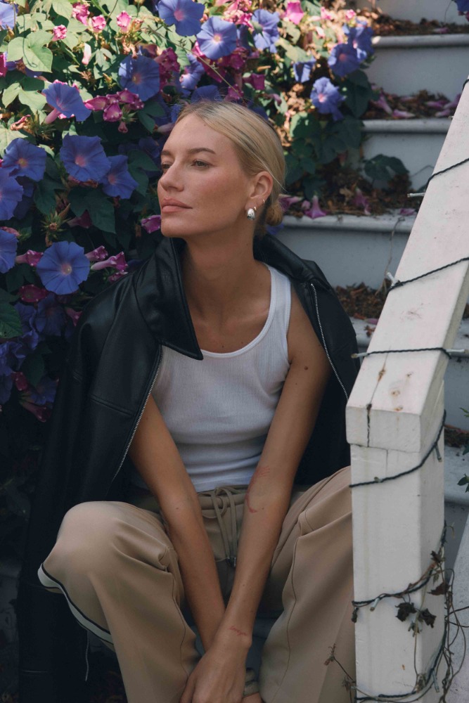 Woman with blonde hair wearing a white top and sweatpants sits on steps with blue flowers in the background, wearing a black jacket.