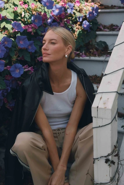 Woman with blonde hair wearing a white top and sweatpants sits on steps with blue flowers in the background, wearing a black jacket.