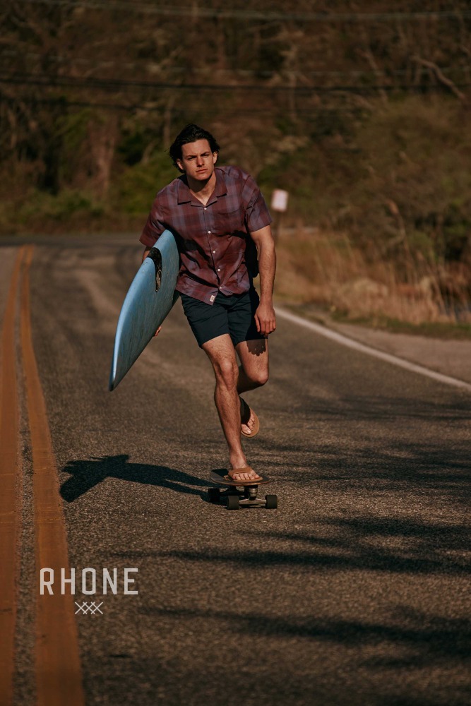 A young man on a skateboard carrying a surfboard on a road. He wears a button-down shirt, dark shorts, and flip-flops.