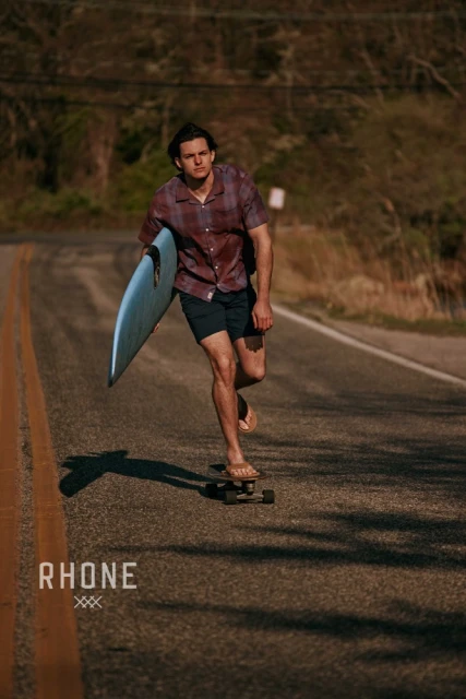 A young man on a skateboard carrying a surfboard on a road. He wears a button-down shirt, dark shorts, and flip-flops.