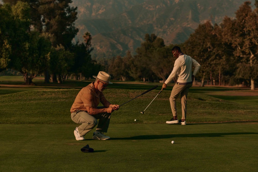 Two men on a golf course, one crouched down and holding a golf club while the other stands preparing to swing, set against a green background.