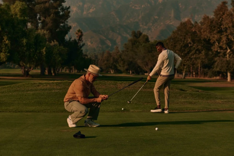Two men on a golf course, one crouched down and holding a golf club while the other stands preparing to swing, set against a green background.