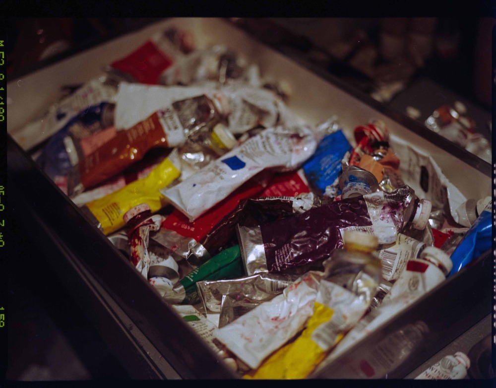 Overhead shot of a bin filled with a disorganized assortment of used paint tubes, showcasing a variety of colors and textures.